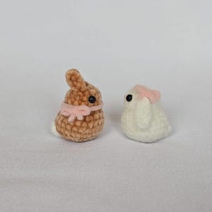 Op de afbeelding: Twee kleine, gehaakte konijnenbeeldjes. E&eacute;n is bruin met een roze strik, de andere is wit met een roze strik. Beide hebben zwarte ogen en staan tegen een witte achtergrond. Deze handgemaakte amigurumi konijntjes zijn perfect voor Pasen.