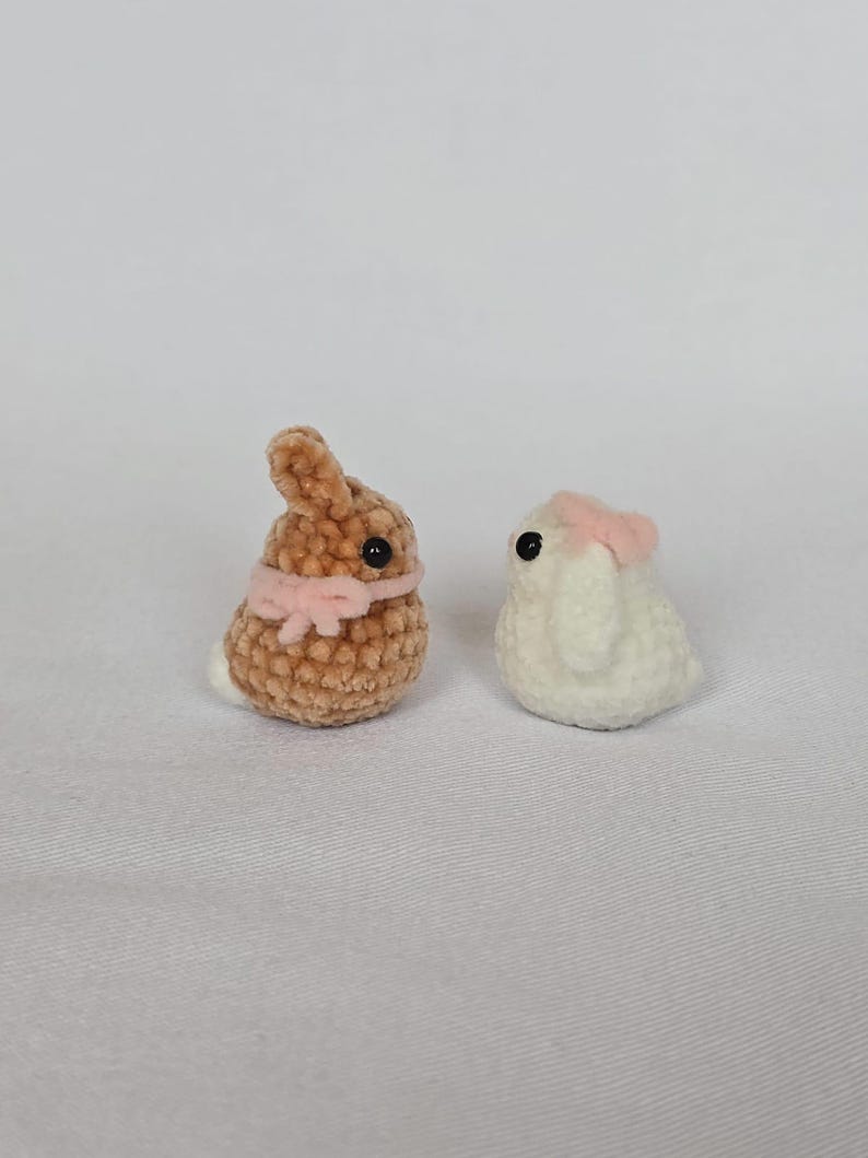 Op de afbeelding: Twee kleine, gehaakte konijnenbeeldjes. E&eacute;n is bruin met een roze strik, de andere is wit met een roze strik. Beide hebben zwarte ogen en staan tegen een witte achtergrond. Deze handgemaakte amigurumi konijntjes zijn perfect voor Pasen.