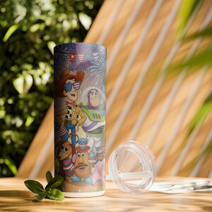May include: A cylindrical tumbler featuring characters from Toy Story, including Woody and Buzz Lightyear, with a festive Fourth of July theme. The design incorporates fireworks, flags, and patriotic colors. A clear lid and straw are included.