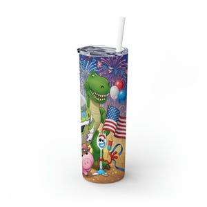 May include: A tall, insulated tumbler with a colorful design featuring characters from the Toy Story movie. The design includes a green dinosaur, a pig, and other characters, along with American flags and fireworks. The tumbler has a silver lid and a white straw.