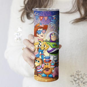 May include: A tall, stainless steel tumbler featuring a patriotic design with cartoon characters, fireworks, and American flags. The tumbler has a silver lid and is held by a person wearing a white sweater.