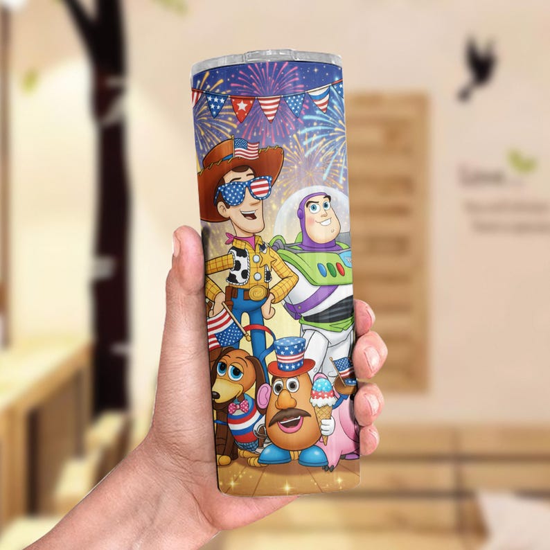 May include: A stainless steel tumbler featuring a colorful illustration of Toy Story characters, including Woody, Buzz Lightyear, and Mr. Potato Head, with a festive fireworks and flag design. The tumbler is held in a hand.