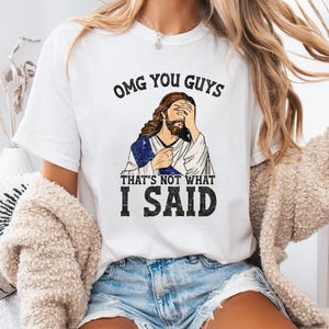 May include: White t-shirt with the text "OMG YOU GUYS" above an illustration of a figure with their face in their hand, and the text "THAT'S NOT WHAT I SAID" below. The figure is wearing a blue and white robe.