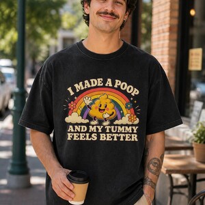 May include: A black short-sleeve t-shirt featuring a graphic of a smiling yellow cartoon character shaped like a poop emoji, a colorful rainbow, and flowers. The text reads I MADE A POOP AND MY TUMMY FEELS BETTER in a retro, cream-colored font.