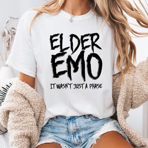 May include: White t-shirt with the text "ELDER EMO" in a bold, black, distressed font. Below the text is the phrase "IT WASN'T JUST A PHASE" in a smaller, black font. The shirt is a casual style.