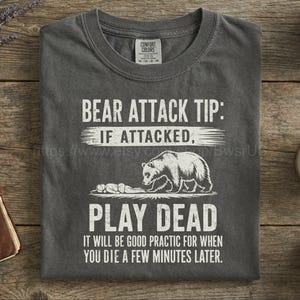 May include: Dark gray t-shirt with the text "BEAR ATTACK TIP: IF ATTACKED, PLAY DEAD" and a graphic of a bear and a person. The text below reads "IT WILL BE GOOD PRACTIC FOR WHEN YOU DIE A FEW MINUTES LATER."