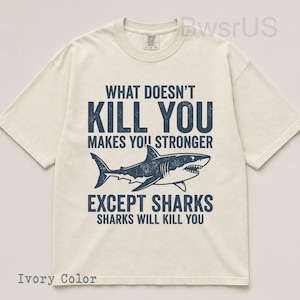 May include: Ivory-colored t-shirt with a navy blue shark graphic and the text "WHAT DOESN'T KILL YOU MAKES YOU STRONGER EXCEPT SHARKS SHARKS WILL KILL YOU." The shirt has a crew neck and short sleeves.