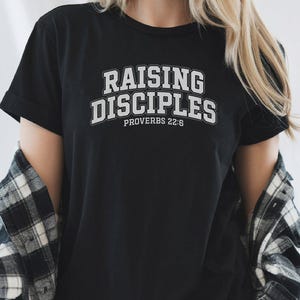 Puede incluir: Camiseta negra de cuello redondo con el texto "RAISING DISCIPLES" en una fuente blanca de estilo universitario. El texto "PROVERBIOS 22:6" está debajo. La camiseta se usa con una camisa a cuadros en blanco y negro.