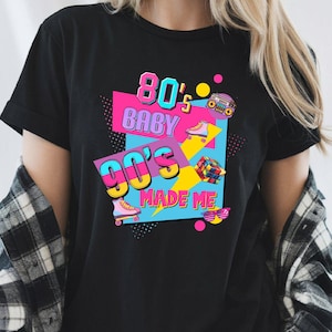 May include: Black t-shirt with a colorful graphic design. The design features the text "80's BABY 90's MADE ME" in vibrant colors, along with images of roller skates, a boombox, a Rubik's Cube, and sunglasses. The shirt has short sleeves.