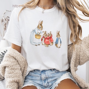 May include: White t-shirt featuring a watercolor illustration of rabbits. The design includes a rabbit wearing a blue dress and holding a basket, with other rabbits in red cloaks and a blue jacket. The text "https://www.etsy.com/shop/BwsrUS" is at the top.
