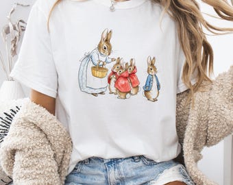 May include: White t-shirt featuring a watercolor illustration of rabbits. The design includes a rabbit wearing a blue dress and holding a basket, with other rabbits in red cloaks and a blue jacket. The text "https://www.etsy.com/shop/BwsrUS" is at the top.
