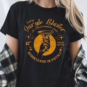 Könnte beinhalten: Schwarzes T-Shirt mit einem verblassten goldenen Aufdruck. Der Aufdruck zeigt die Worte "Enjoy Gargle Blaster" und "Resistance is Futile" um ein Cocktailglas, das von einer Hand gehalten wird. Der Text "Drink and Don't Panic" ist ebenfalls enthalten.