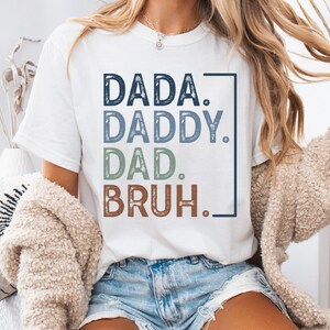 May include: White t-shirt featuring the words "DADA. DADDY. DAD. BRUH." in a vertical layout. The text is in shades of blue, green, and brown, within a dark blue rectangle. The shirt is a relaxed fit.