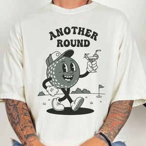 May include: Cream-colored t-shirt featuring a cartoon golf ball character with a cap, holding a cocktail. The text "ANOTHER ROUND" is arched above the character. The design is in black and white.