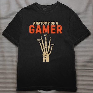 Op de afbeelding: Zwart t-shirt met de tekst "ANATOMY OF A GAMER" in oranje letters. Daaronder een skelet hand grafiek met de letters "A", "W/S", en "D" boven de vingers, en "SHIFT/CTRL" en "SPACE" aan de zijkanten.