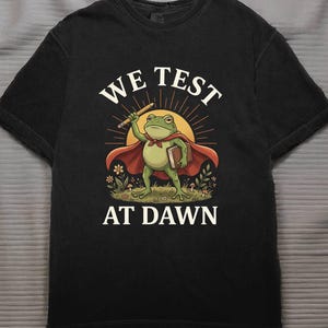 May include: Black t-shirt featuring a cartoon frog superhero with a pencil and book, wearing a red cape. The text "WE TEST AT DAWN" is arched above and below the frog illustration, set against a sunburst background.