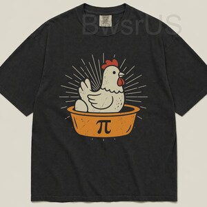 May include: Dark gray t-shirt featuring a cartoon chicken in an orange pie dish with the pi symbol. The chicken has a red comb and is surrounded by radiating lines. The shirt is a relaxed fit.