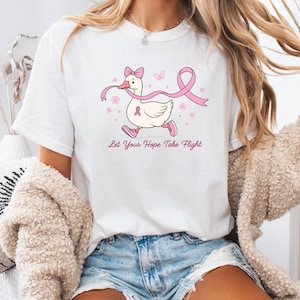 May include: White t-shirt featuring a cartoon duck wearing a pink ribbon and running. The duck has a pink bow and pink shoes. Pink ribbon and flowers are also on the shirt. The text "Let Your Hope Take Flight" is below the duck.
