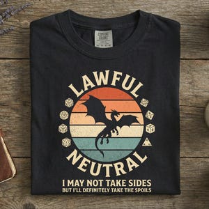 May include: Black t-shirt with a circular graphic featuring a dragon silhouette, dice, and the words "LAWFUL NEUTRAL." Below the graphic, the text reads "I MAY NOT TAKE SIDES BUT I'LL DEFINITELY TAKE THE SPOILS."