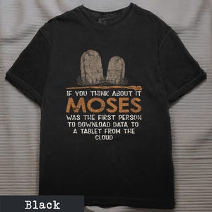 May include: Black t-shirt with a humorous graphic. The design features two tombstone illustrations and the text: "IF YOU THINK ABOUT IT MOSES WAS THE FIRST PERSON TO DOWNLOAD DATA TO A TABLET FROM THE CLOUD."