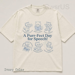 May include: Ivory-colored t-shirt featuring a playful design with blue illustrations of kittens engaged in various activities. The text "A Purr-Fect Day for Speech!" is prominently displayed, along with related phrases. The shirt is a casual wear item.
