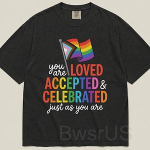 May include: Black t-shirt with a rainbow flag graphic and the words "you are LOVED ACCEPTED & CELEBRATED just as you are" in colorful text. The shirt is a casual style.