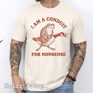 May include: Ivory-colored t-shirt with a whimsical design. The graphic features a frog riding a goose, with the text "I AM A CONDUIT FOR NONSENSE" in red. The shirt is a crew neck and short-sleeved.