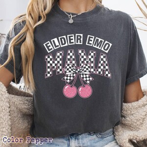 May include: Dark gray t-shirt with the text "ELDER EMO MAMA" in white and pink. The word "MAMA" is in a checkered pattern, with a bow and two pink cherries below. The shirt is a casual style.