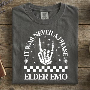 May include: Dark gray t-shirt with the text "IT WAS NEVER A PHASE ELDER EMO" in white. The design includes a skeleton hand making a rock and roll gesture, lightning bolts, and a checkered pattern.