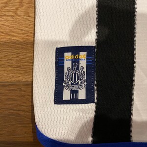 May include: Close-up of a white and black soccer jersey with a blue trim. The jersey features a navy blue patch with the Adidas logo and the words "OFFICIAL REPLICA" above a crest and the words "NEWCASTLE UNITED".