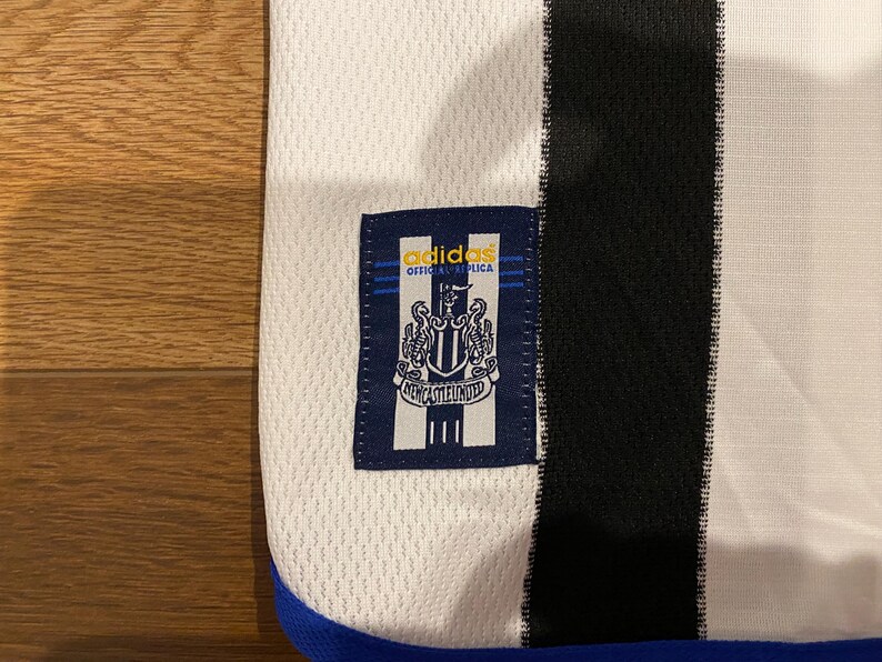 May include: Close-up of a white and black soccer jersey with a blue trim. The jersey features a navy blue patch with the Adidas logo and the words "OFFICIAL REPLICA" above a crest and the words "NEWCASTLE UNITED".