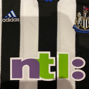 May include: A black and white striped jersey with the Adidas logo and a Newcastle United crest. The jersey also features the word "ntl" in purple and green letters.