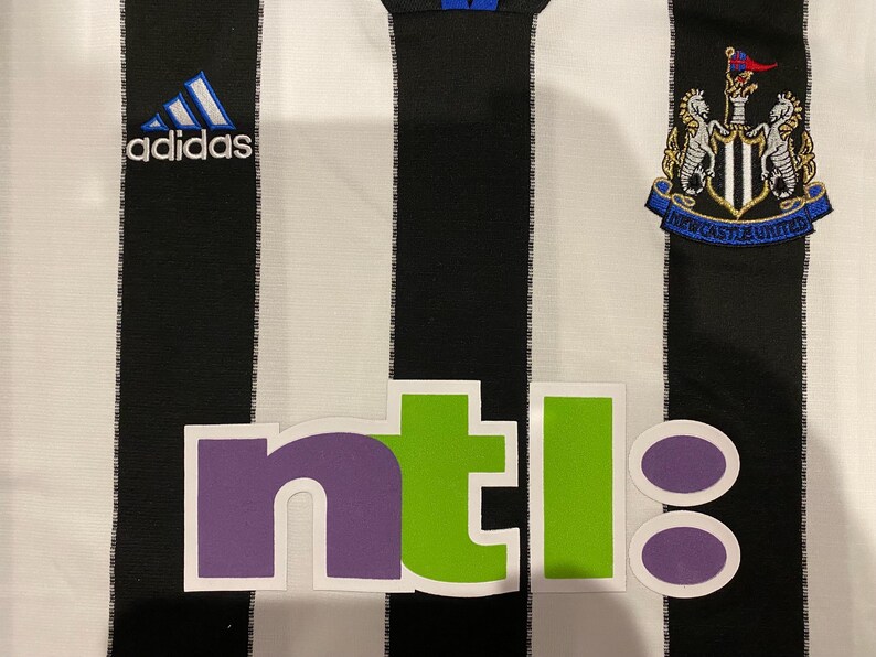 May include: A black and white striped jersey with the Adidas logo and a Newcastle United crest. The jersey also features the word "ntl" in purple and green letters.