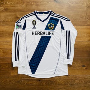 May include: White long-sleeved soccer jersey with navy blue accents and a v-neck collar. Features the text "HERBALIFE" and the LA Galaxy team logo. Includes the Adidas logo and MLS patch.