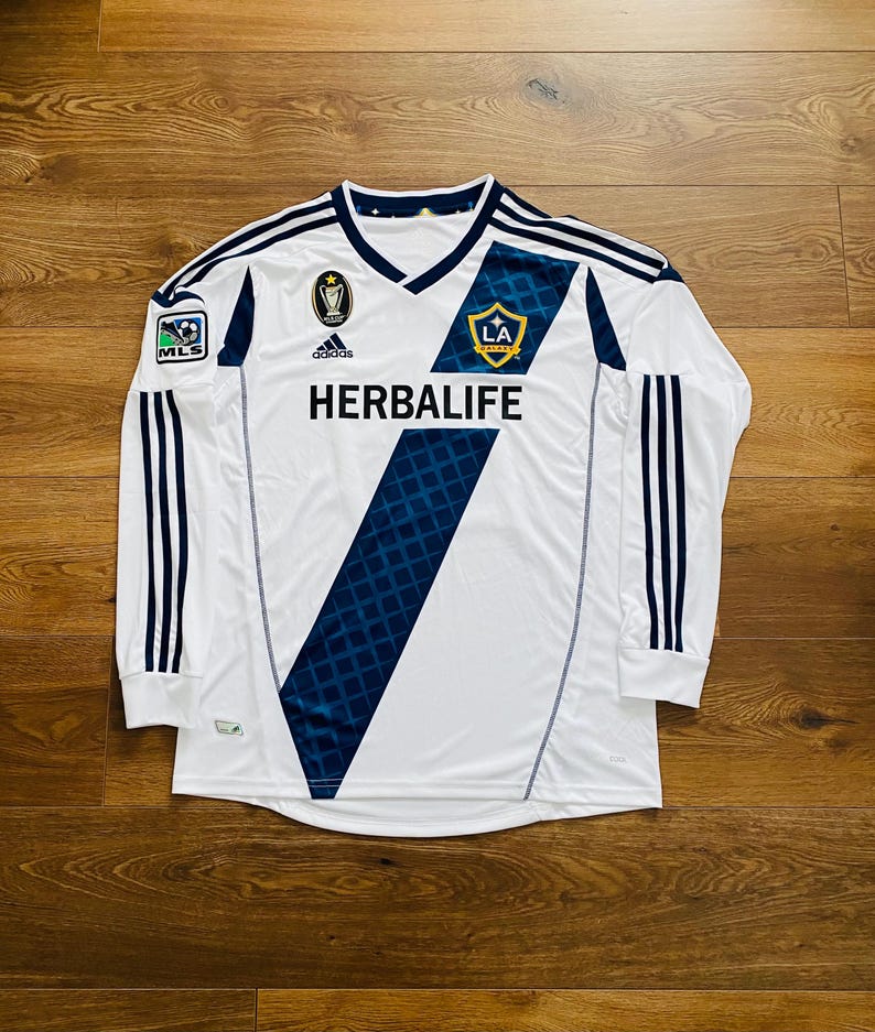 May include: White long-sleeved soccer jersey with navy blue accents and a v-neck collar. Features the text "HERBALIFE" and the LA Galaxy team logo. Includes the Adidas logo and MLS patch.