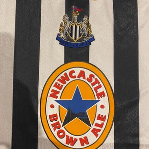 Może przedstawiać: Zbliżenie koszulki piłkarskiej Newcastle United w czarno-białe paski. Logo Adidas jest haftowane na złoto. Koszulka posiada herb drużyny i okrągłe logo z napisem "Newcastle Brown Ale".