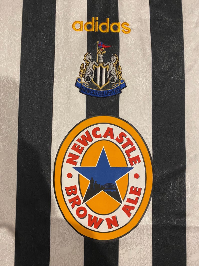 Może przedstawiać: Zbliżenie koszulki piłkarskiej Newcastle United w czarno-białe paski. Logo Adidas jest haftowane na złoto. Koszulka posiada herb drużyny i okrągłe logo z napisem "Newcastle Brown Ale".