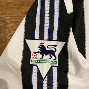 May include: Close-up of a black and white striped jersey with a blue trim. The jersey features a Premier League logo with a lion and a soccer ball. The jersey is made of a lightweight material.