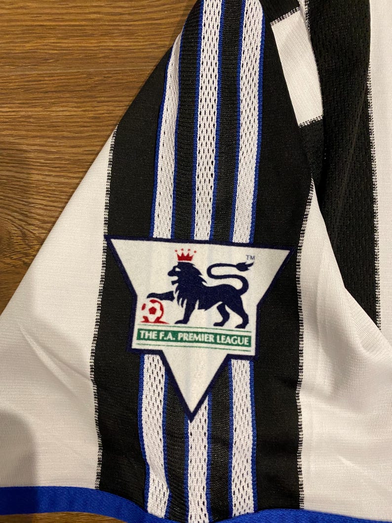 May include: Close-up of a black and white striped jersey with a blue trim. The jersey features a Premier League logo with a lion and a soccer ball. The jersey is made of a lightweight material.