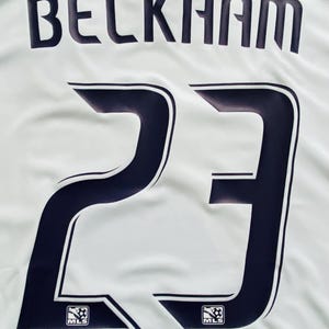 May include: White jersey with the name "BECKHAM" in navy blue block letters above the number "23". The number is navy blue with a white outline. The MLS logo is at the bottom of the number.