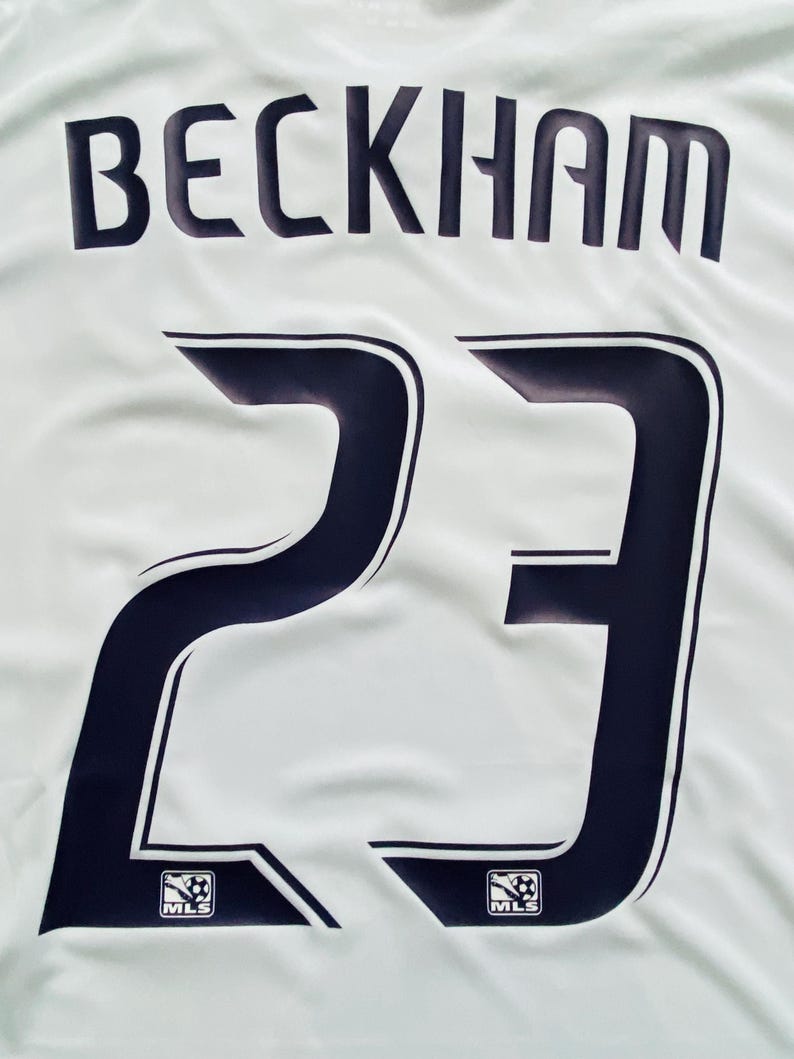 May include: White jersey with the name "BECKHAM" in navy blue block letters above the number "23". The number is navy blue with a white outline. The MLS logo is at the bottom of the number.