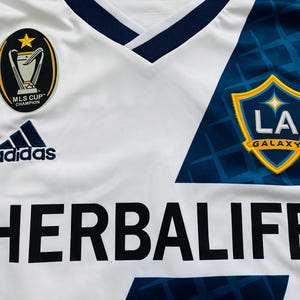 May include: White soccer jersey with blue and gold accents. Features the Adidas logo, MLS Cup Champion patch, and the LA Galaxy team logo. The jersey also displays the Herbalife sponsor logo in large black letters.