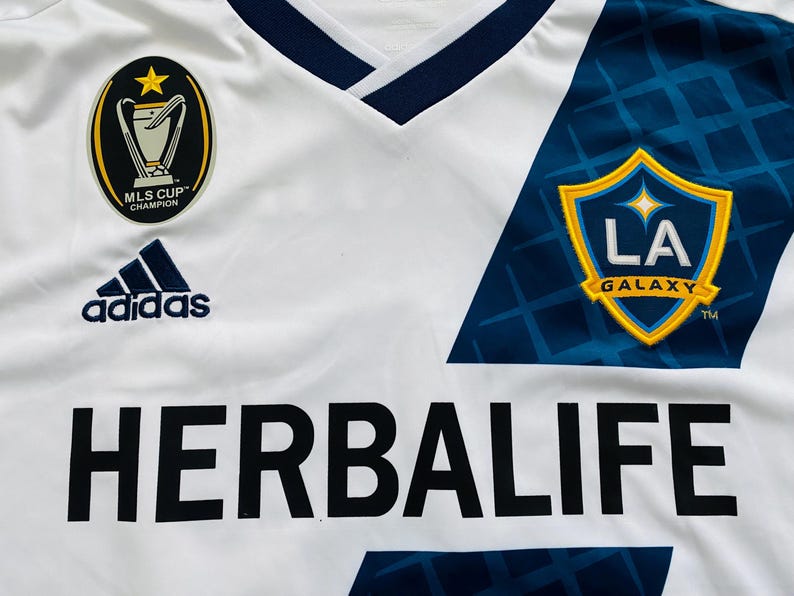 May include: White soccer jersey with blue and gold accents. Features the Adidas logo, MLS Cup Champion patch, and the LA Galaxy team logo. The jersey also displays the Herbalife sponsor logo in large black letters.