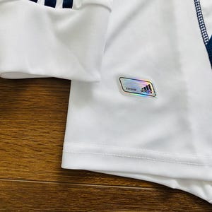 May include: Close-up of a white athletic jersey with a blue geometric pattern on the side. The sleeve cuff has three blue stripes. A holographic Adidas label is visible.