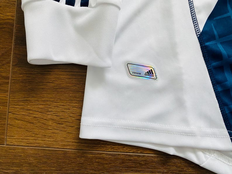 May include: Close-up of a white athletic jersey with a blue geometric pattern on the side. The sleeve cuff has three blue stripes. A holographic Adidas label is visible.