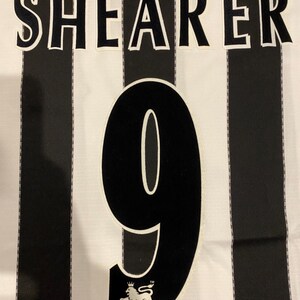 May include: A black and white striped jersey with the name "SHEARER" across the top and the number "9" in the center. The Premier League logo is at the bottom. The jersey is made of a lightweight fabric.