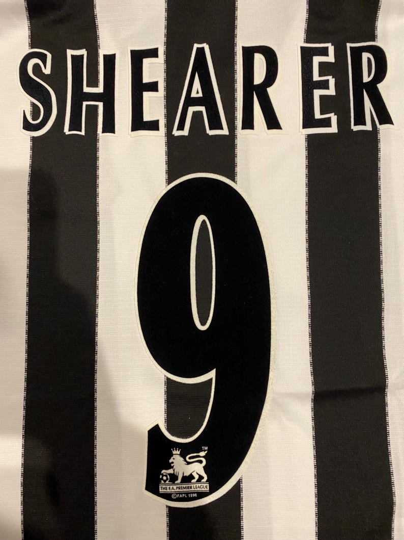 May include: A black and white striped jersey with the name "SHEARER" across the top and the number "9" in the center. The Premier League logo is at the bottom. The jersey is made of a lightweight fabric.
