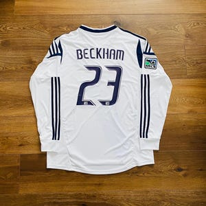 May include: White long-sleeved soccer jersey with the name "BECKHAM" and the number "23" in navy blue. Features navy blue stripes on the sleeves and a MLS logo patch. The jersey is displayed on a wooden surface.