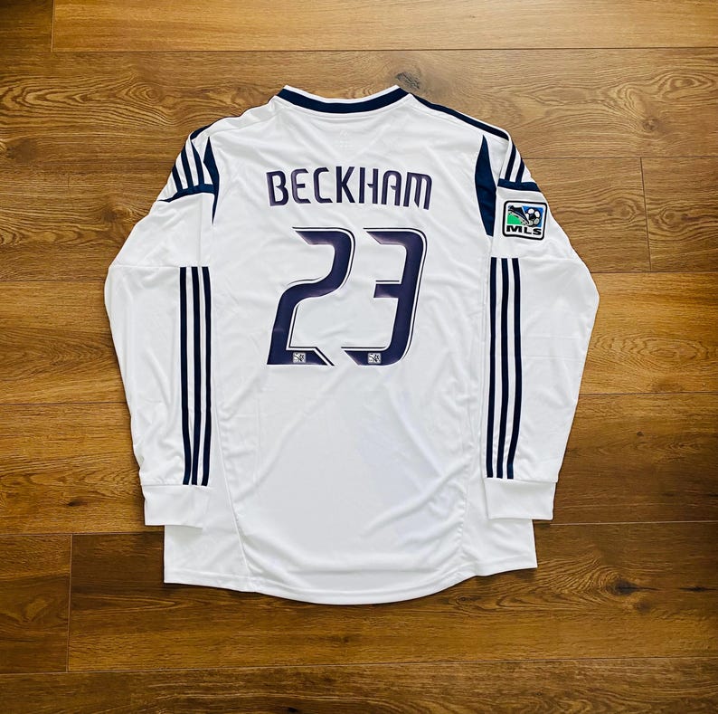 May include: White long-sleeved soccer jersey with the name "BECKHAM" and the number "23" in navy blue. Features navy blue stripes on the sleeves and a MLS logo patch. The jersey is displayed on a wooden surface.