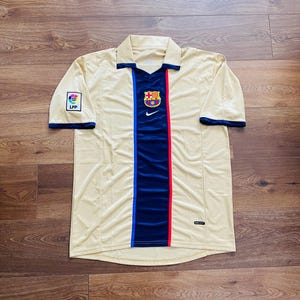 May include: A tan football shirt with a navy blue and red vertical stripe. The shirt features a collared neckline with navy trim and short sleeves. The team emblem is on the chest, and the LFP logo is on the left sleeve.